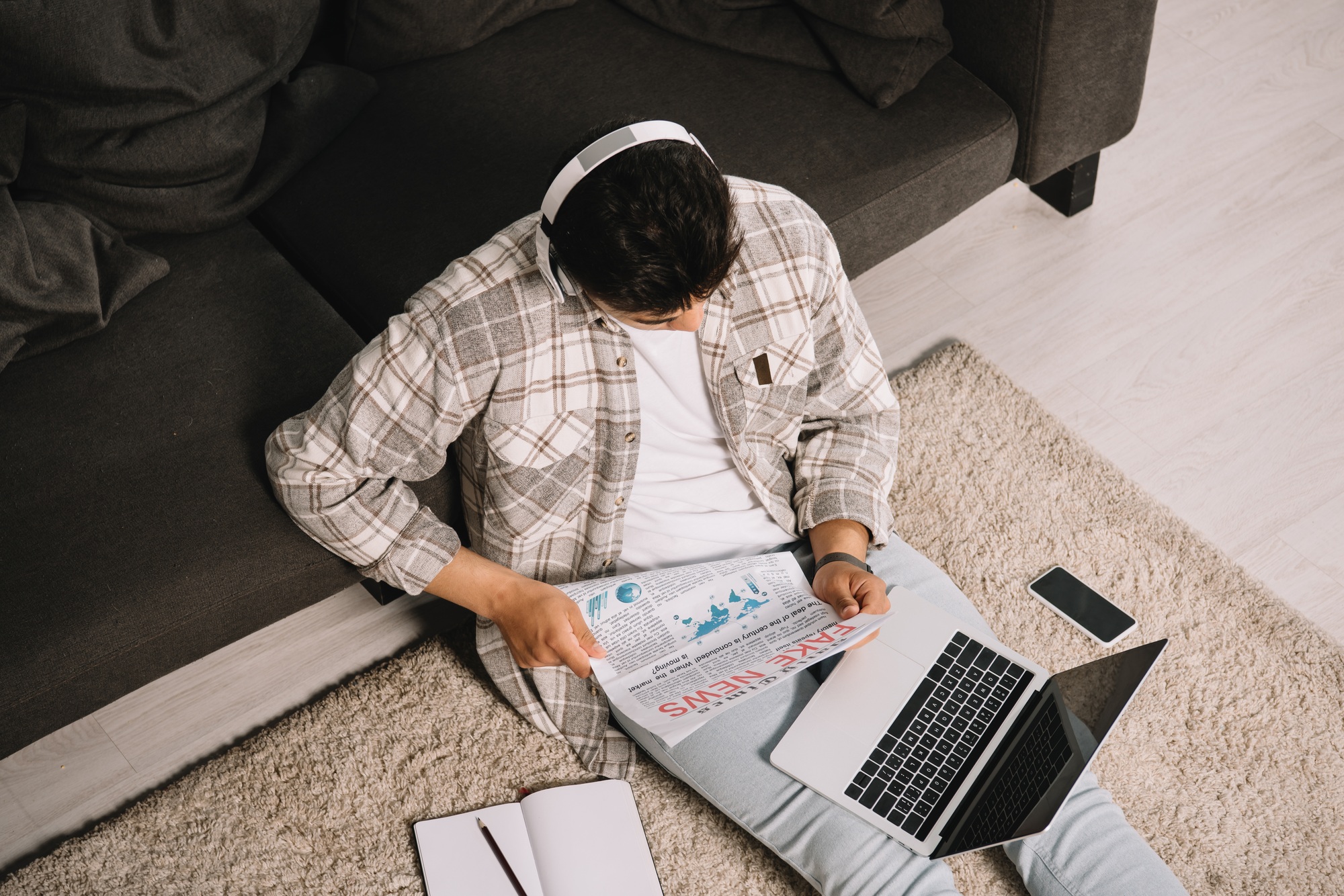 overhead view of young man reading fake news newspaper while sitting on floor with laptop